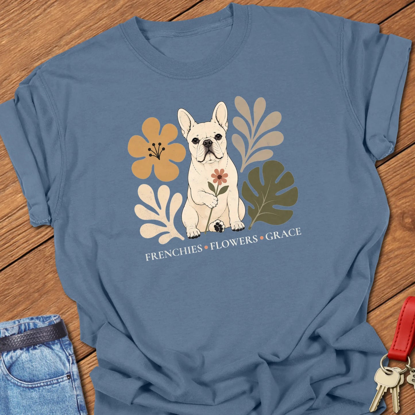 Cream French Bulldog holding a flower surrounded by modern botanical artwork with the phrase "Frenchies • Flowers • Grace" printed on a Comfort Colors® garment-dyed t-shirt in blue Jean color.