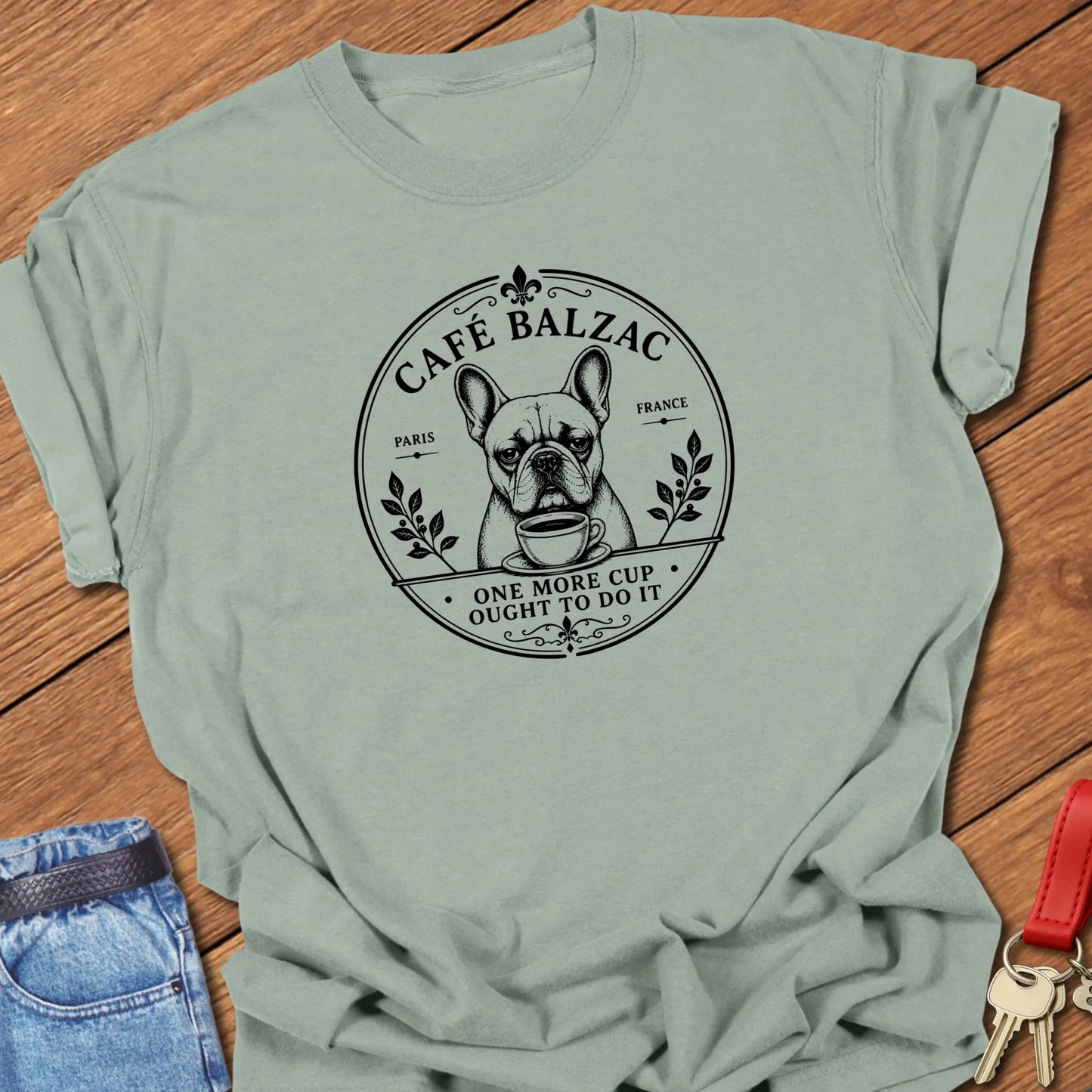 Café Balzac French Bulldog graphic t-shirt featuring "One More Cup Ought To Do It" slogan with vintage Paris café artwork inspired by literary coffee culture in Bay color.