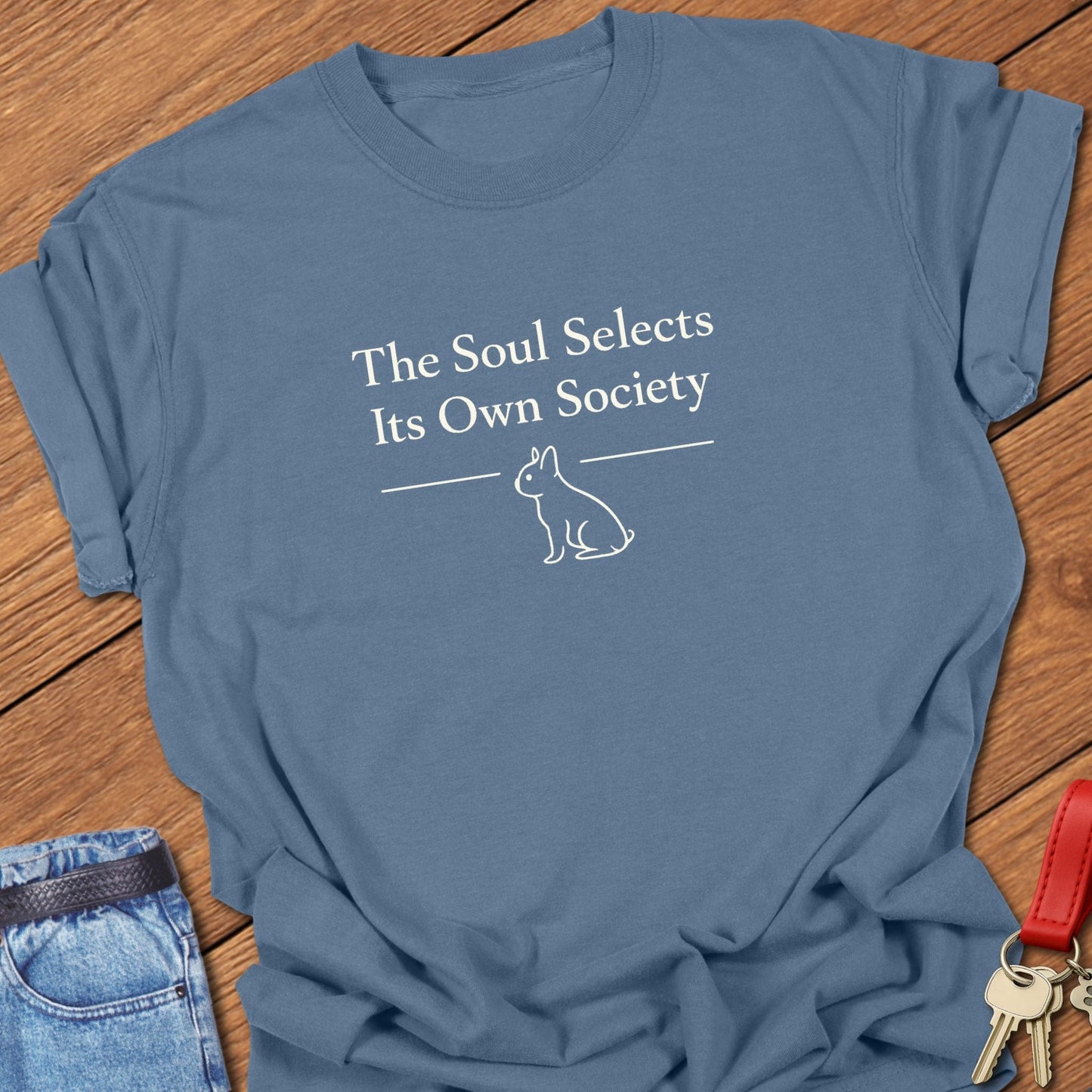 Ivory serif text reading “The Soul Selects Its Own Society” by Emily Dickinson above a minimalist French bulldog silhouette printed on a Comfort Colors blue jean t shirt.
