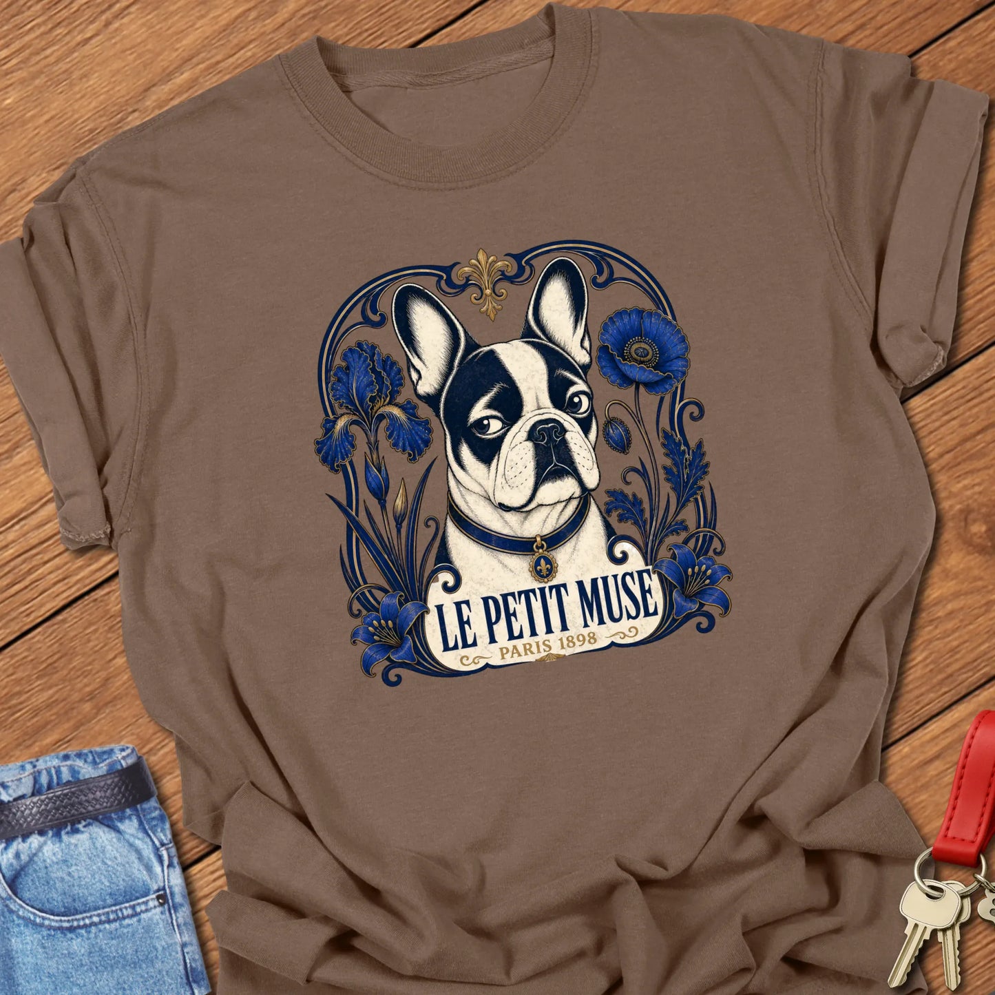 Art Nouveau-inspired French Bulldog portrait surrounded by cobalt-blue floral ornamentation with the title "Le Petit Muse — Paris 1898" printed on a Comfort Colors® garment-dyed t-shirt in Espresso color.